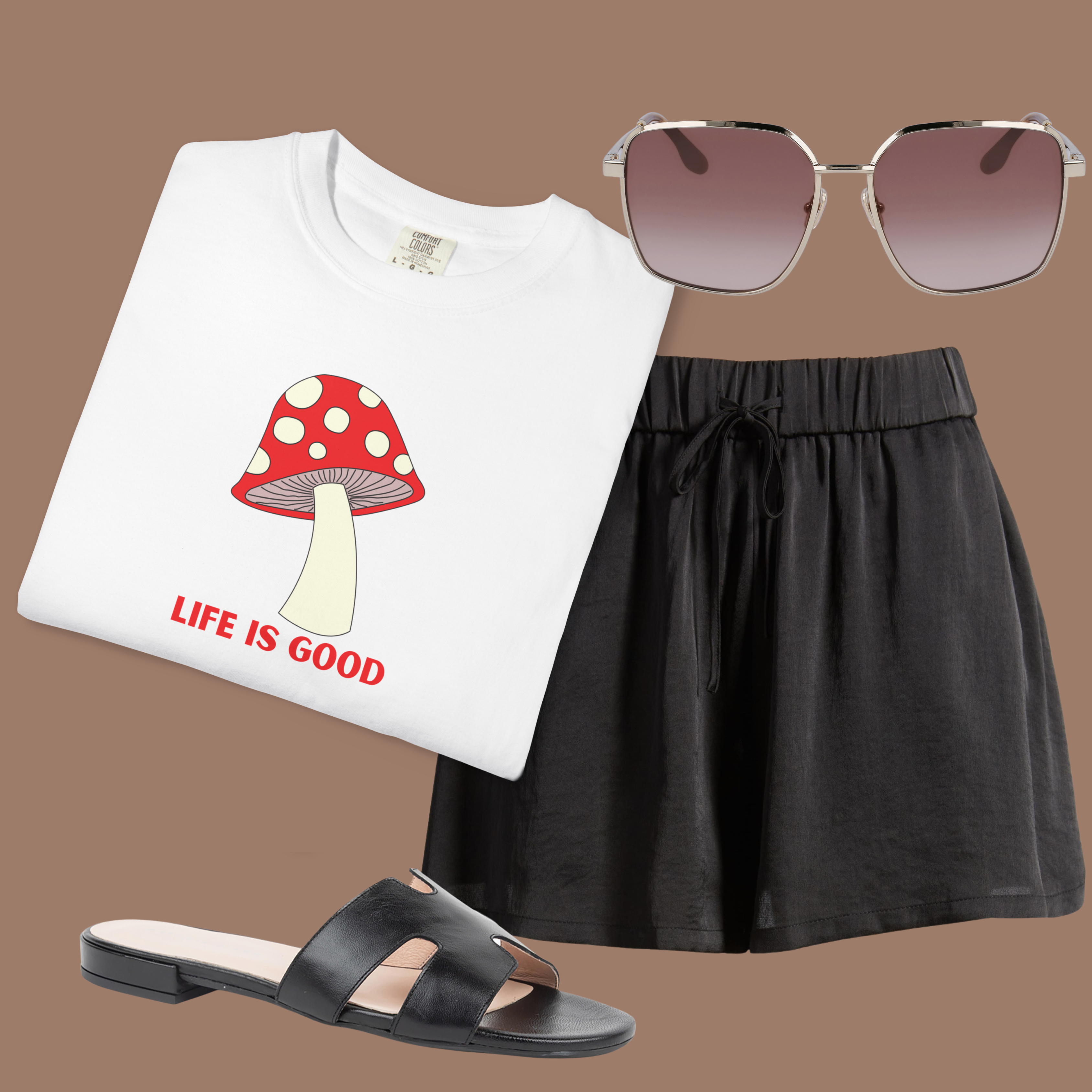 White Comfort Colors tee with red mushroom and “Life Is Good” text, styled with black shorts, sandals, and sunglasses for a sunny, casual look.